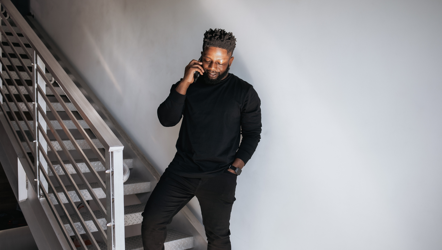 A man in dark clothing and glasses standing by a metal staircase, talking on a phone