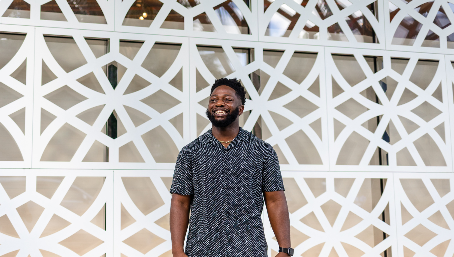 A smiling man in a patterned short-sleeve shirt standing in front of a white geometric lattice wall