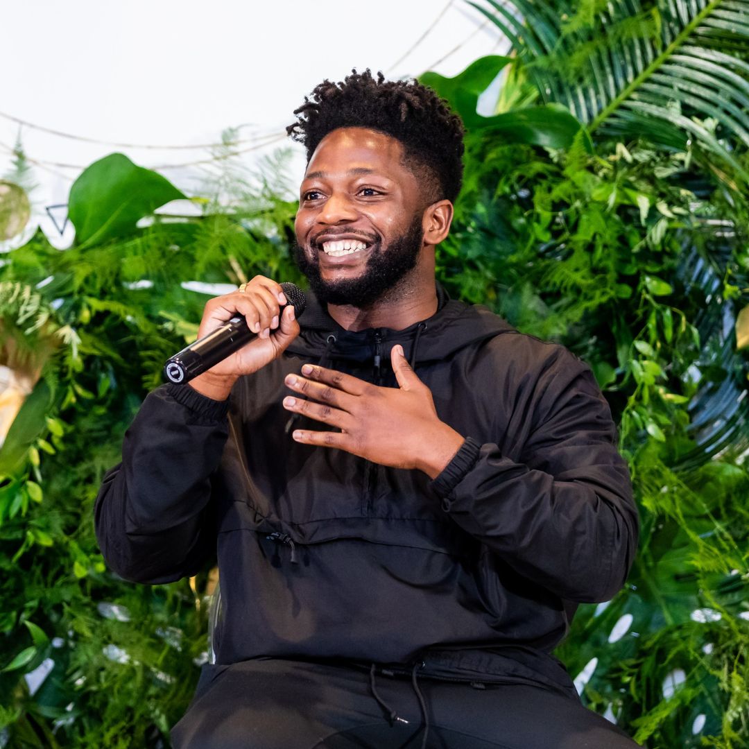 A smiling man in a black hoodie speaking into a microphone in front of green plants